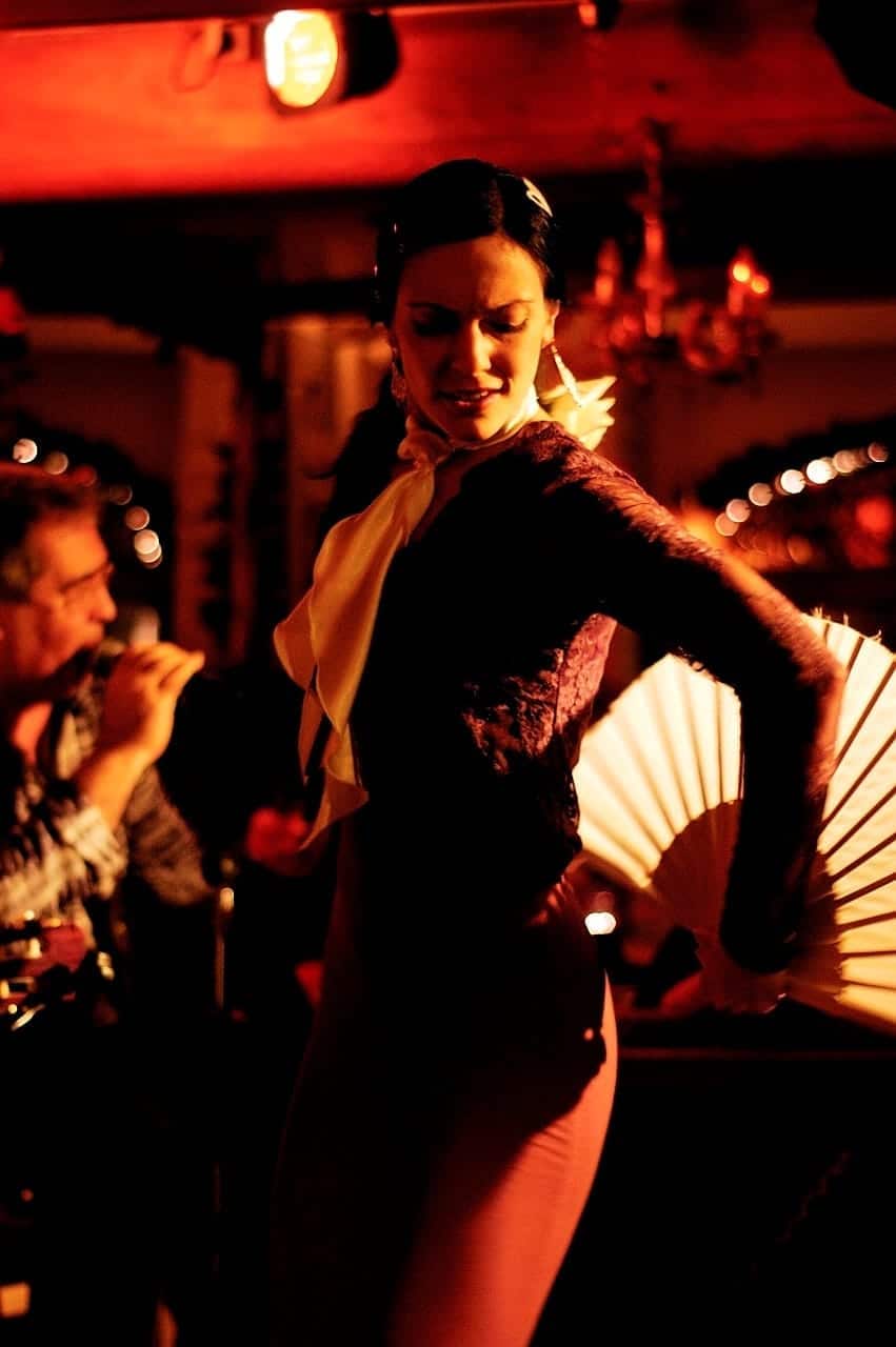 Bailaora con un abanico blanco y blusa de encaje negra en un tablao, con un músico al fondo