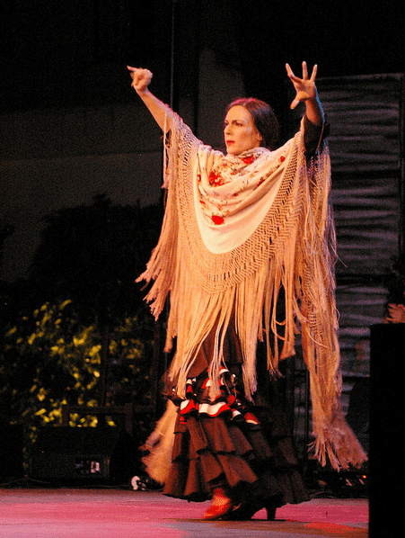 Mónica Vázquez bailando flamenco en un escenario con un mantón de flecos y los brazos en alto